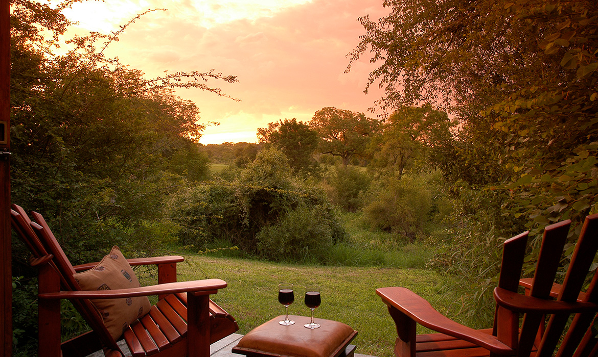 Duas cadeiras de madeira de frente para a savana. Ao fundo vegetação verde e o amanhecer em tons de amarelo e laranja se mistura.
Entre as cadeiras um puff serve duas taças de vinho tinto.