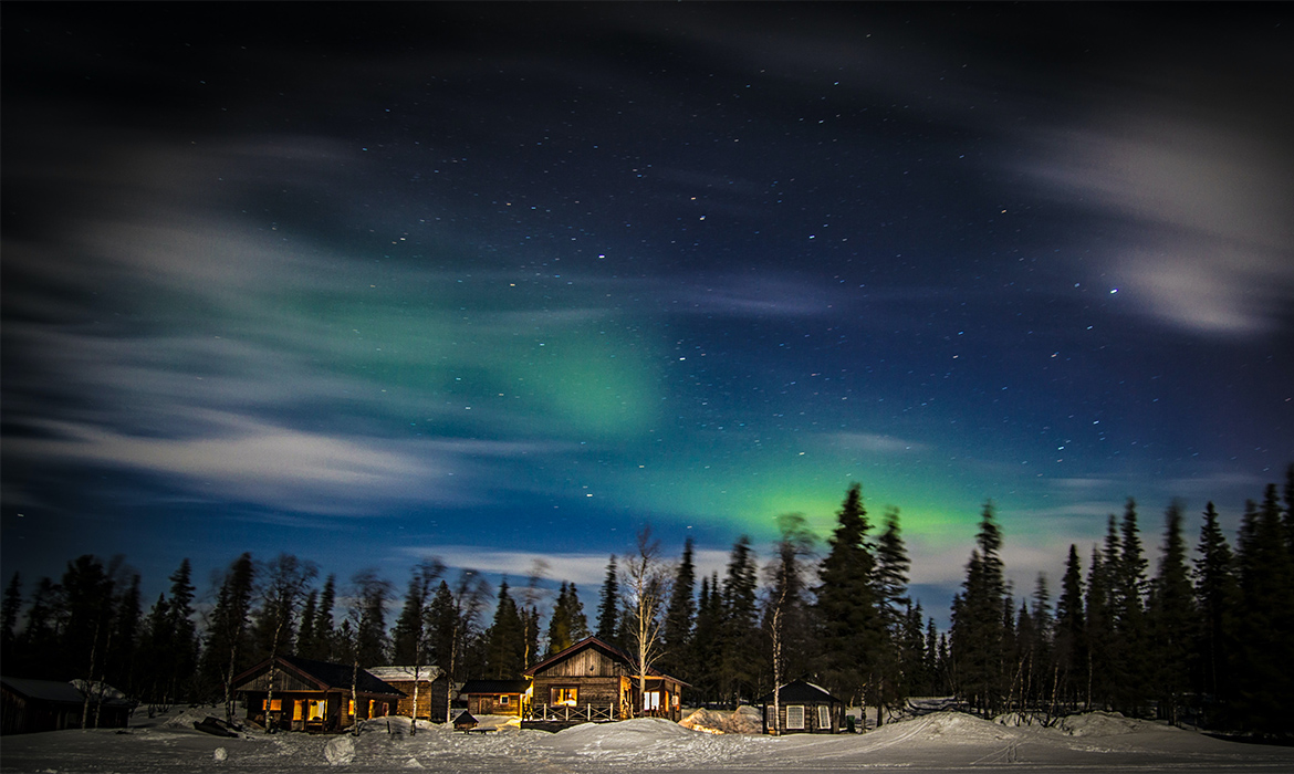 pequenos focos de luzes verdes no céu estrelado. A paisagem compõe a sombra de pinheiros e pequenas casas de madeira. Chão coberto de neve